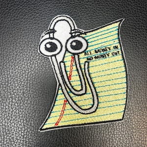 May include: An embroidered patch featuring a white paperclip character with large eyes and a worried expression, positioned over a yellow lined paper background. The text ALL MONEY IN NO MONEY OUT is stitched in black thread on the paper.