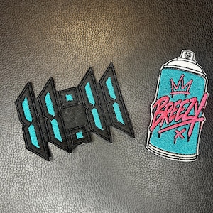 May include: Two embroidered patches: one featuring the numbers 11:11 in a digital clock style with black borders and bright teal centers, and another depicting a teal spray paint can with a pink crown and the word BREEZY written in pink.
