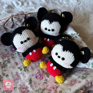 May include: Three handmade crochet Mickey Mouse keychains. Each keychain features a black head with large ears, a white face, red shorts, and yellow shoes. The keychains are displayed on a floral patterned fabric.