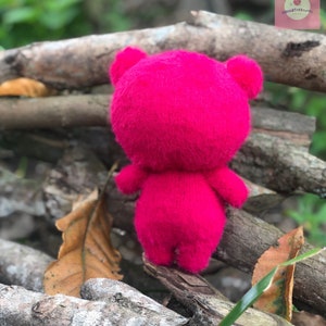 Puede incluir: Un oso de peluche rosa brillante hecho a mano, visto desde atr&aacute;s, con orejas y extremidades visibles. El juguete de peluche se coloca sobre ramas de madera natural y follaje, creando un contraste de texturas y colores.