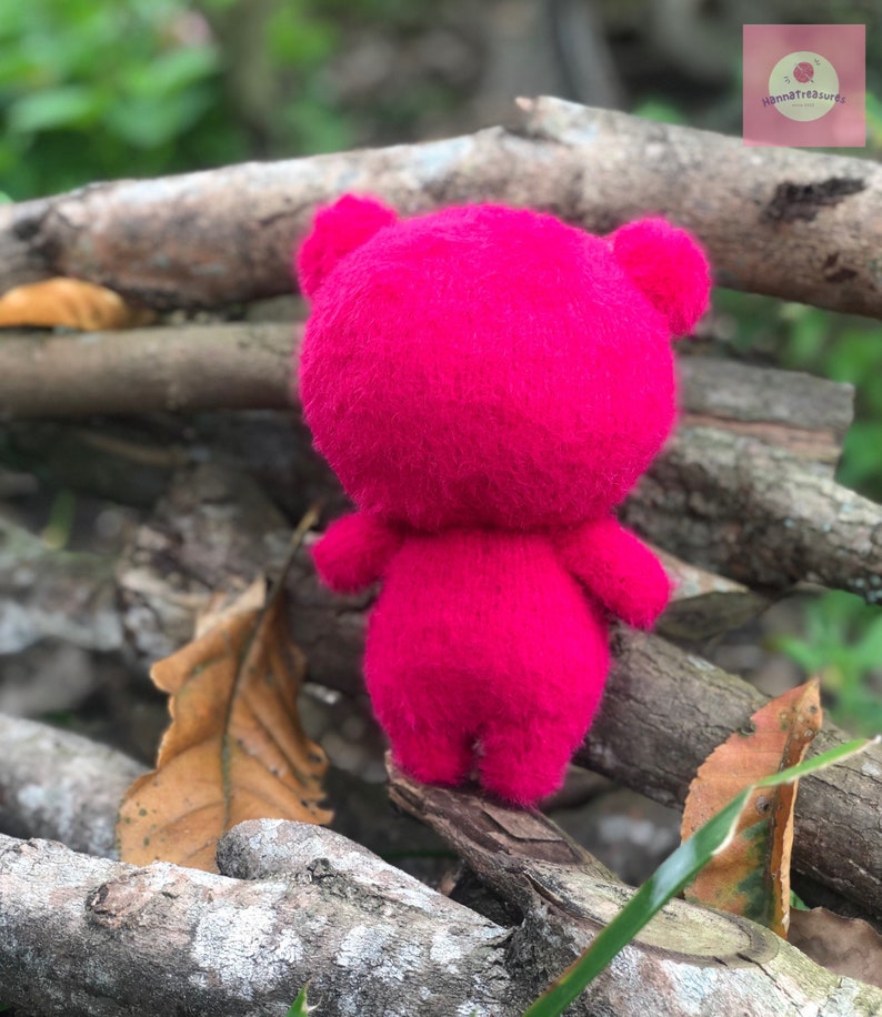 Puede incluir: Un oso de peluche rosa brillante hecho a mano, visto desde atr&aacute;s, con orejas y extremidades visibles. El juguete de peluche se coloca sobre ramas de madera natural y follaje, creando un contraste de texturas y colores.