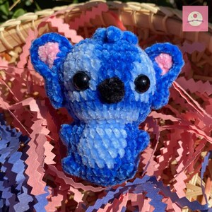 Puede incluir: Un juguete de peluche azul hecho a mano con apariencia de koala. El juguete presenta grandes ojos negros, una nariz negra y detalles rosas en el interior de las orejas. El peluche es de material suave y texturizado, sobre un fondo de papel rosa y azul.