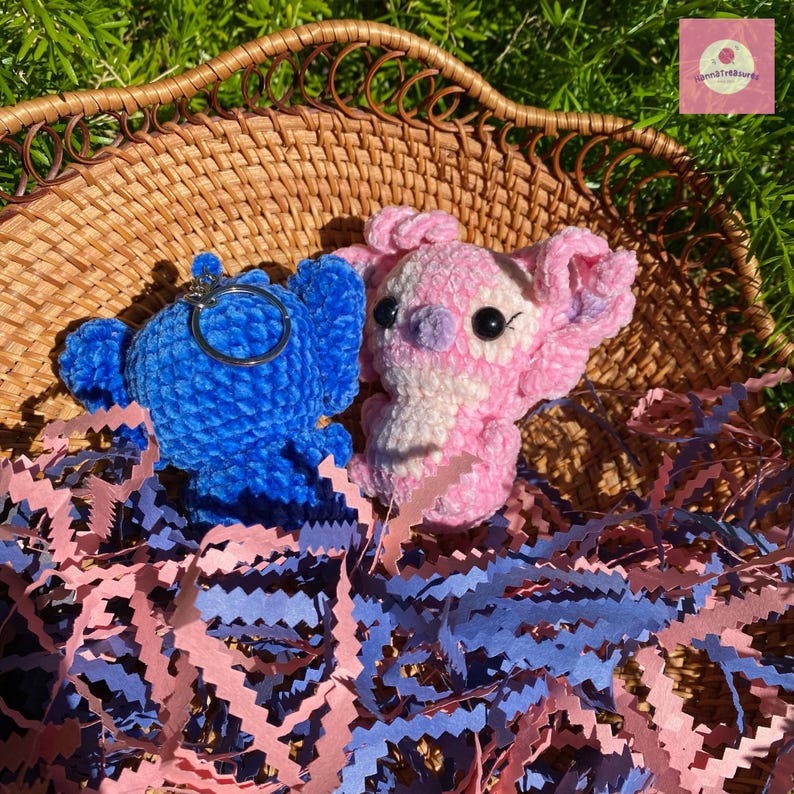 Puede incluir: Dos figuras de peluche de ganchillo, una azul con un llavero y otra rosa y blanca, est&aacute;n en una cesta de mimbre. Papel triturado rosa y azul rodea las figuras, creando una escena colorida y juguetona.