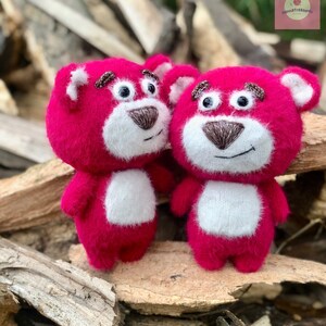Puede incluir: Dos osos de peluche rosas con vientres y caras blancas. Los osos tienen narices y cejas marrones bordadas y ojos negros. Los osos est&aacute;n de pie sobre un trozo de madera.