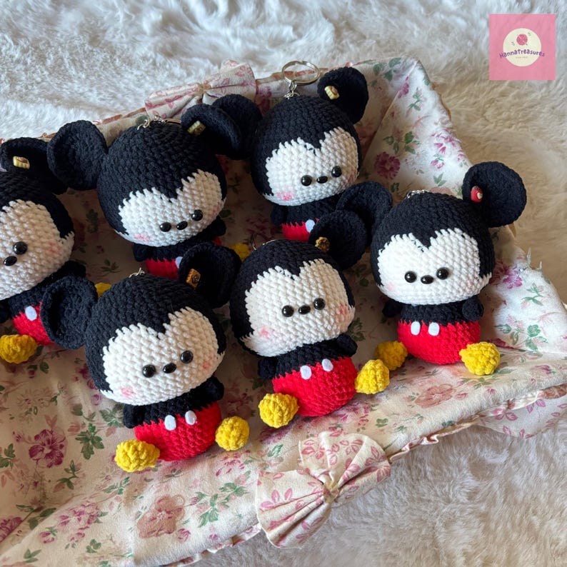 May include: A collection of handmade crochet Mickey Mouse keychains. Each keychain features a black head and ears, a white face, red shorts, and yellow shoes. The keychains are displayed in a floral-lined basket.