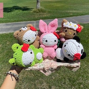 May include: A collection of six crocheted plush toys featuring Hello Kitty in different animal costumes. The toys are pink, green, brown, and white with red accents. The toys are sitting on a floral patterned fabric.