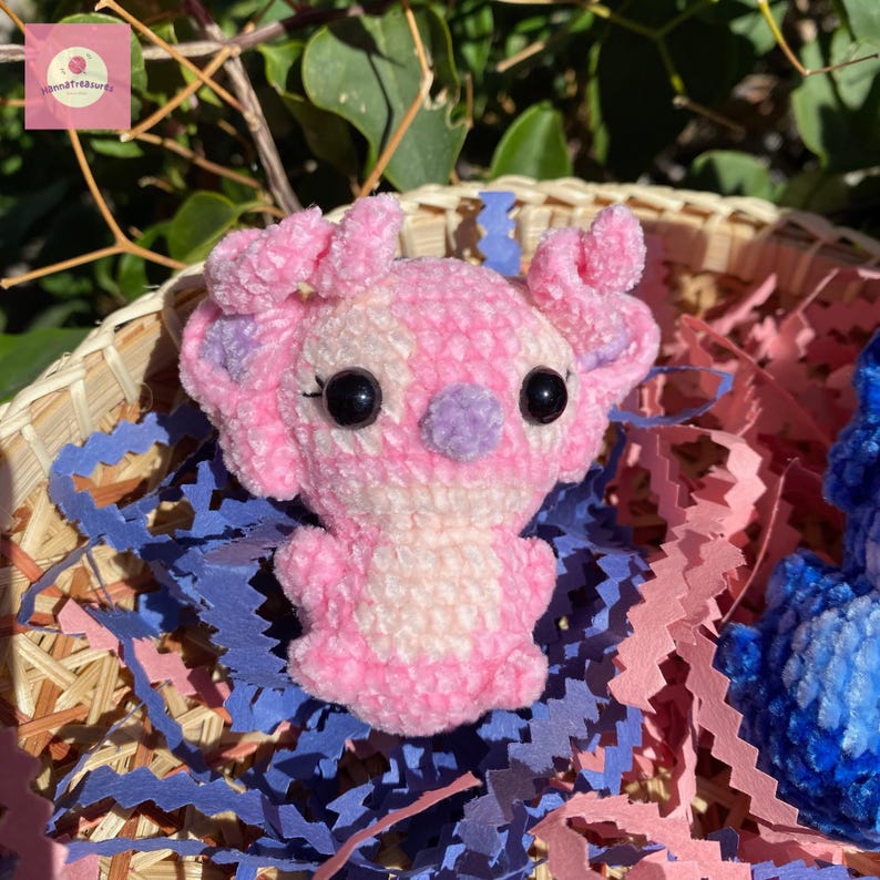 Puede incluir: Una criatura de ganchillo rosa y blanco hecha a mano, con grandes ojos negros y detalles morados. El peluche se exhibe en una cesta tejida con papel rosa y azul. Un art&iacute;culo de artesan&iacute;a hecho a mano.