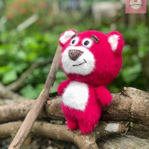 Puede incluir: Un oso de peluche hecho a mano, de color rosa vibrante, con detalles blancos en el pecho y las orejas. El oso tiene una nariz marr&oacute;n y detalles bordados. El juguete est&aacute; colocado sobre ramas de madera, con un fondo verde borroso.