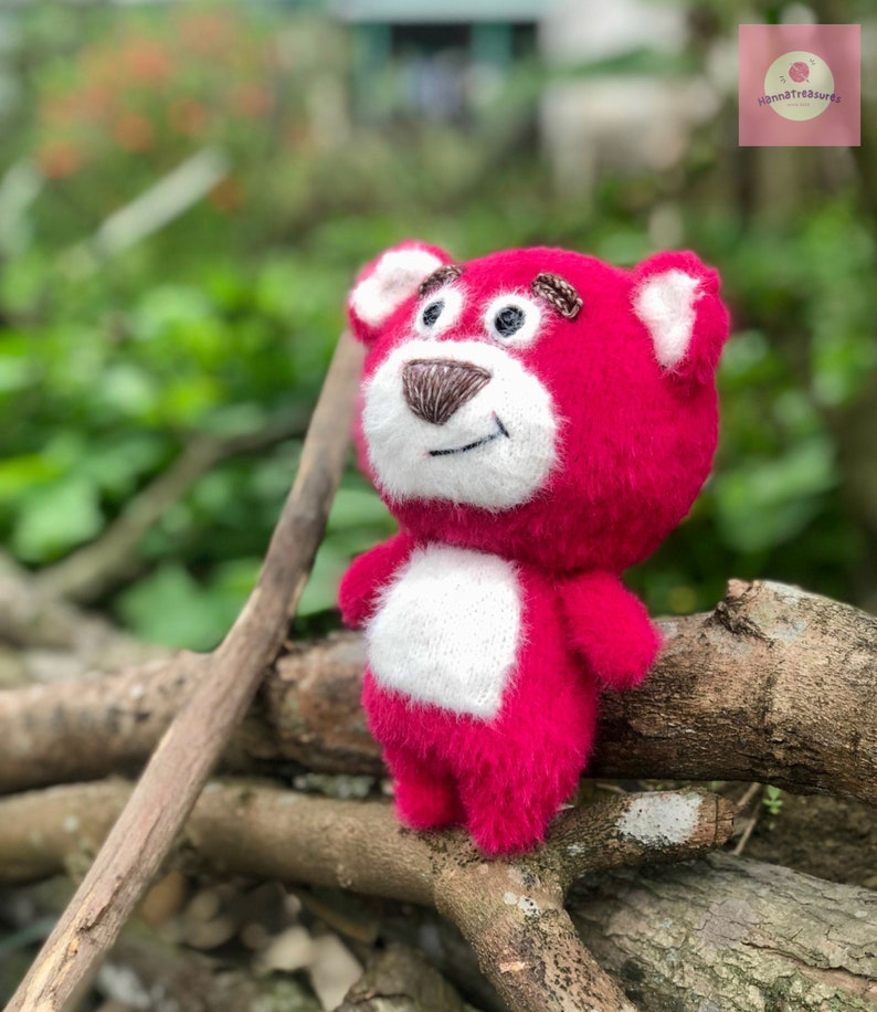 Puede incluir: Un oso de peluche hecho a mano, de color rosa vibrante, con detalles blancos en el pecho y las orejas. El oso tiene una nariz marr&oacute;n y detalles bordados. El juguete est&aacute; colocado sobre ramas de madera, con un fondo verde borroso.