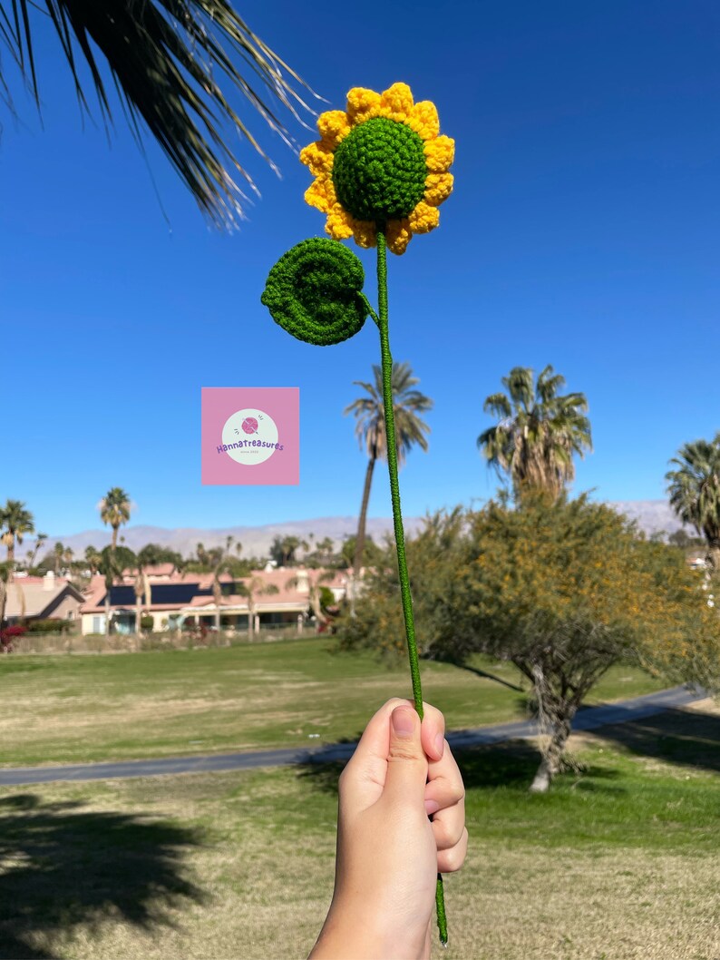 May include: A crocheted sunflower with a green center and yellow petals. The sunflower is on a green stem and has a green leaf.