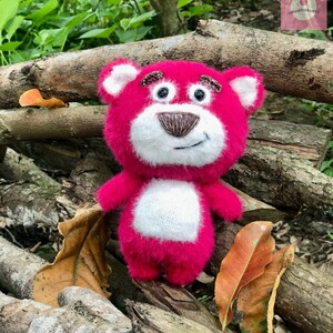 Puede incluir: Un oso de peluche rosa hecho a mano con detalles blancos en la cara y el vientre. El oso tiene rasgos bordados, incluyendo una nariz y cejas marrones, sobre un fondo natural de madera y hojas.