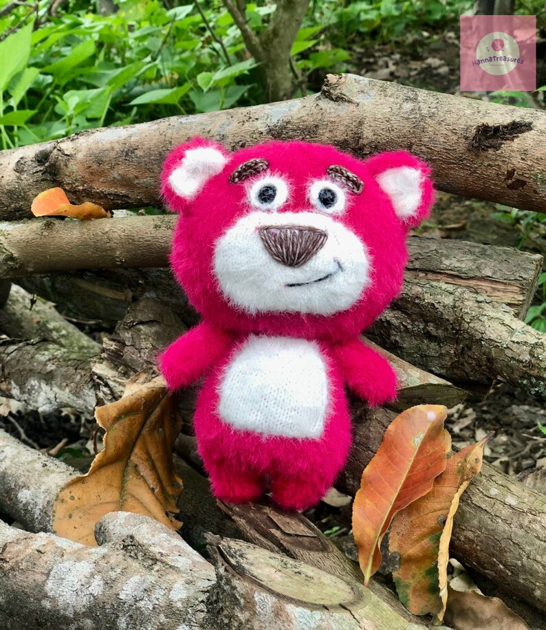 Puede incluir: Un oso de peluche rosa hecho a mano con detalles blancos en la cara y el vientre. El oso tiene rasgos bordados, incluyendo una nariz y cejas marrones, sobre un fondo natural de madera y hojas.