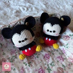May include: Two handmade crochet Mickey Mouse plushies. Each plushie features a black head with large ears, a white face, a red body with white buttons, and yellow feet. The plushies are displayed on a floral patterned fabric.