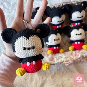 May include: Crocheted Mickey Mouse keychain with black ears, a white face, and a red body. The keychain has yellow feet and a silver keyring. Several other Mickey Mouse keychains are visible in the background.