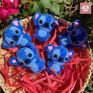 Puede incluir: Colecci&oacute;n de koalas azules de ganchillo con ojos negros y orejas rosas, dispuestos en una cesta de mimbre con papel rojo. Un koala tiene un llavero. Los koalas est&aacute;n en varias poses, algunos tumbados, otros de pie.