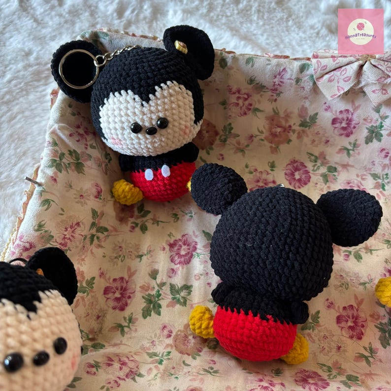 May include: Crocheted Mickey Mouse keychains. The keychains feature black heads with white faces, black ears, red shorts, and yellow shoes. The keychains are placed on a floral patterned fabric.