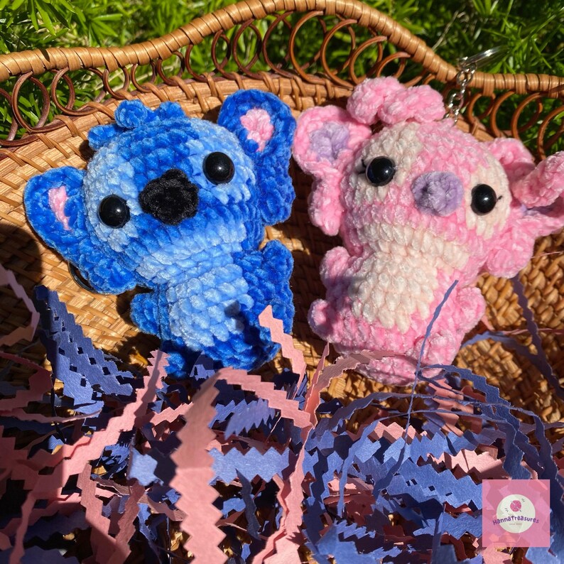 Puede incluir: Dos llaveros de peluche con forma de personajes de dibujos animados. Uno es azul con detalles rosas, el otro es rosa y blanco. Ambos tienen ojos negros y est&aacute;n hechos de un material suave y texturizado. Se encuentran sobre papel triturado dentro de una cesta tejida.