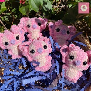 Puede incluir: Un conjunto de llaveros de koala tejidos en rosa y blanco. Los koalas tienen ojos negros y detalles morados en las orejas y la nariz. Est&aacute;n colocados sobre papel azul triturado en una cesta tejida.