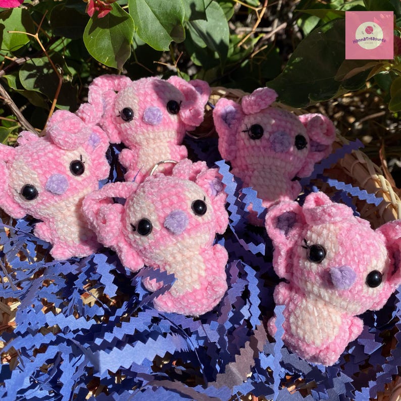 Puede incluir: Un conjunto de llaveros de koala tejidos en rosa y blanco. Los koalas tienen ojos negros y detalles morados en las orejas y la nariz. Est&aacute;n colocados sobre papel azul triturado en una cesta tejida.