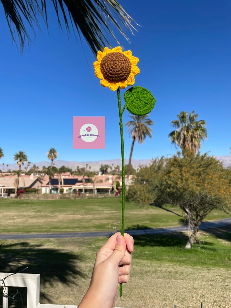 May include: A crocheted sunflower with a brown center and yellow petals, a green leaf, and a green stem. The sunflower is held in a hand against a background of a blue sky and green grass.