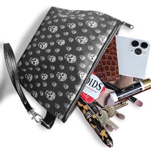May include: A black cosmetic bag with a skull pattern, open to reveal a phone, makeup, keys, and a pack of mints. The bag has a wrist strap and a zip closure. The skull pattern is white on a black background.