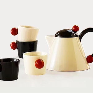 May include: A cream-colored teapot with a black lid and handle, accompanied by black and cream-colored cups. Each cup and the teapot feature a red sphere as a handle or accent. A modern tea set.