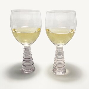 May include: Two clear wine glasses filled with a light yellow liquid. The glasses have a ribbed stem and a rounded bowl. The glasses are on a white surface.