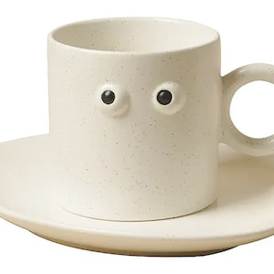 May include: Cream-colored ceramic coffee cup and saucer set. The cup features two black, round eye-shaped details and a circular handle. The set has a speckled texture and a minimalist design, suitable for serving coffee or tea.