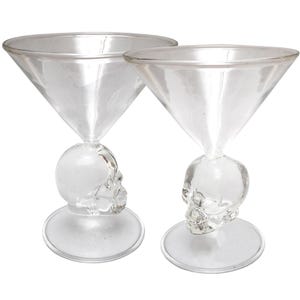 May include: Two clear glass martini glasses featuring a sculpted skull design as the stem. Each glass has a wide, conical bowl, a detailed human skull shaped base support, and a circular foot for stability. The glassware is transparent and colourless.