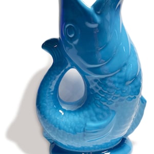 May include: A turquoise ceramic pitcher in the shape of a fish. The pitcher has a wide open mouth, a tail-shaped handle, and a round base. The fish has textured scales and a glossy finish.