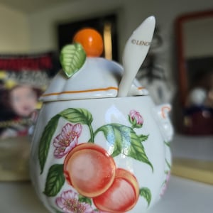 May include: A white porcelain sugar bowl featuring a painted peach and blossom motif. The lid is topped with a sculpted orange fruit and green leaf. A matching white porcelain spoon with the text LENOX is tucked into the lid opening.