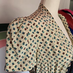 Könnte beinhalten: Cremefarbenes Kleid mit einem kleinen, sich wiederholenden Muster aus blauen und roten herzförmigen Designs. Das Kleid hat kurze Ärmel, einen V-Ausschnitt und eine taillierte Taille. Weiße Knöpfe zieren die Vorderseite, und der Stoff wirkt leicht.