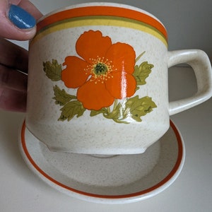 May include: Cream-colored ceramic mug and saucer set. The mug features a large orange flower design with green leaves. The rim has orange and yellow stripes. The saucer has a matching orange rim. The set is likely vintage.