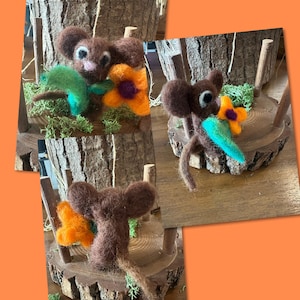 Peut inclure: Figurine de souris en feutre faite à la main avec de grandes oreilles, tenant une feuille verte et une fleur orange. La souris est marron avec un nez rose et des yeux blancs. La figurine est posée sur une base en bois avec de la mousse.