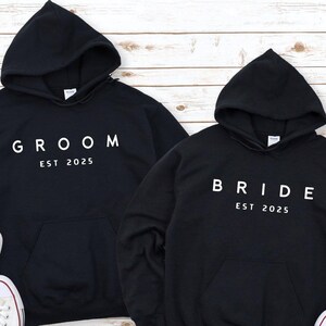 May include: Two black hooded sweatshirts with white text. One sweatshirt says "Groom Est 2025" and the other says "Bride Est 2025".