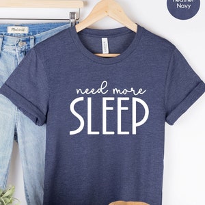 May include: A heather navy blue t-shirt with the text "need more SLEEP" in white. The shirt is displayed on a hanger with a pair of jeans, a potted plant, a hat, and white sneakers.