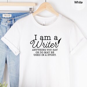 May include: White t-shirt with the text "I am a Writer. Anything you say or do may be used in a story."  The shirt is displayed on a hanger with a pair of blue jeans, a hat, and a plant.