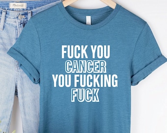 May include: A teal t-shirt with the text "FUCK YOU CANCER YOU FUCKING FUCK" in white. The shirt is displayed with light blue jeans, white trainers, and a straw hat. A small potted plant is also visible.