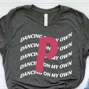 May include: A dark gray t-shirt with the text "Dancing on my own" repeated in white lettering. A large pink "P" is centered on the shirt.