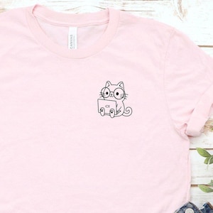 May include: A light pink t-shirt with a small black and white cat graphic, paired with denim shorts and white sneakers. The t-shirt features a crew neck and short sleeves. The sneakers have white laces and red accents.