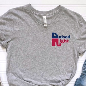 May include: Gray t-shirt with a red, white, and blue elephant graphic and the text "raised right". The shirt is paired with blue jeans and white sneakers.