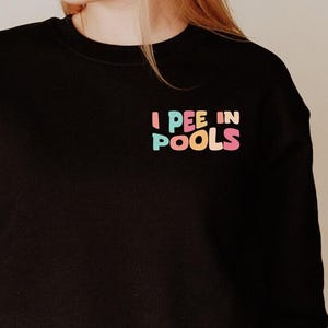 May include: Black sweatshirt with the text "I PEE IN POOLS" in pastel colors. The sweatshirt is a crew neck style and is made of a soft material. The text is printed on the upper left chest area.