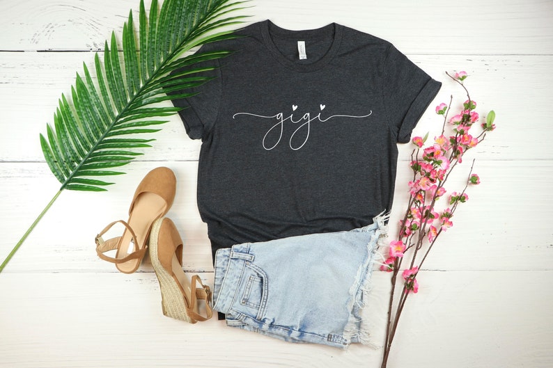 May include: A dark gray t-shirt with the word "gigi" written in white cursive with two small hearts above the word. The t-shirt is laying on a white wooden surface with a pair of brown sandals, a pair of light blue denim shorts, and a sprig of pink flowers.