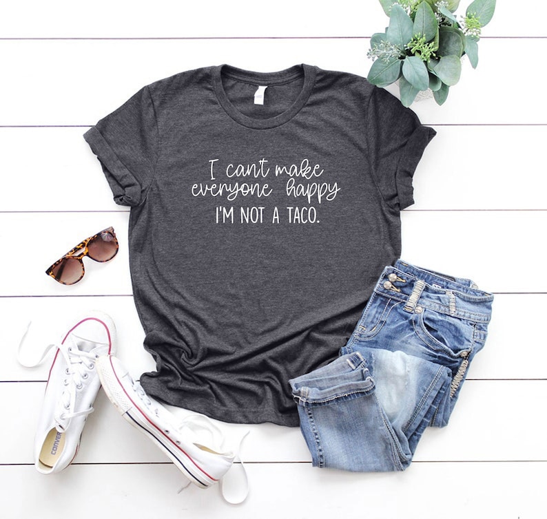 Puede incluir: Una camiseta gris oscuro con el texto "I can't make everyone happy I'm not a taco." impreso en blanco en la parte delantera. La camiseta est&aacute; colocada sobre una superficie de madera blanca con unas gafas de sol, unas zapatillas blancas y unos vaqueros azules.