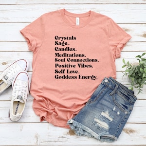 May include: A peach-colored t-shirt with the text "Crystals Sage. Candles. Meditations. Soul Connections. Positive Vibes. Self Love. Goddess Energy." printed in black.