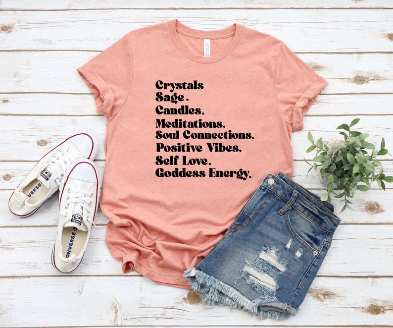 May include: A peach-colored t-shirt with the text "Crystals Sage. Candles. Meditations. Soul Connections. Positive Vibes. Self Love. Goddess Energy." printed in black.