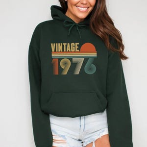 Pode incluir: Um moletom verde escuro com um bolso frontal e um capuz com cord&atilde;o. A frente do moletom apresenta a palavra "VINTAGE" acima do ano "1976" em um esquema de cores retr&ocirc;, com um gr&aacute;fico de p&ocirc;r do sol.