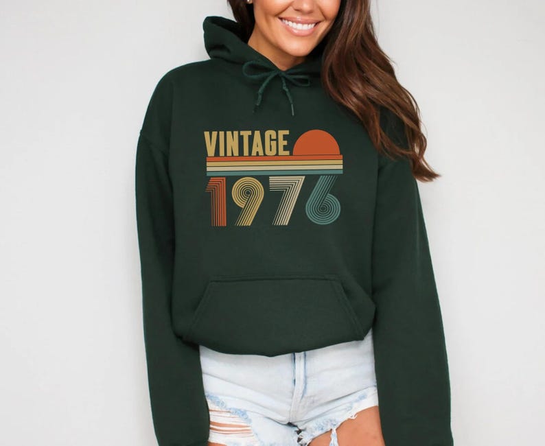 Pode incluir: Um moletom verde escuro com um bolso frontal e um capuz com cord&atilde;o. A frente do moletom apresenta a palavra "VINTAGE" acima do ano "1976" em um esquema de cores retr&ocirc;, com um gr&aacute;fico de p&ocirc;r do sol.