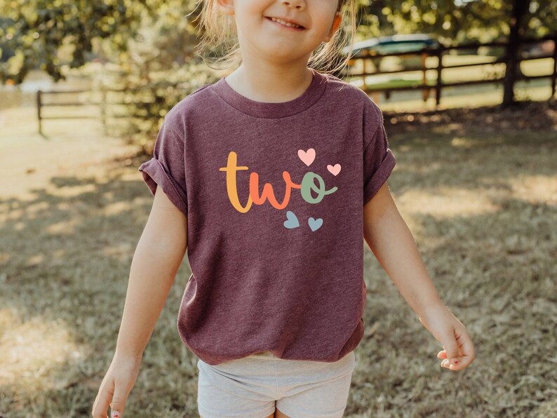 May include: A child wearing a purple t-shirt with the word "two" written in rainbow colors and surrounded by colorful hearts.