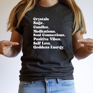 May include: A dark grey t-shirt with white text that reads "Crystals Sage. Candles. Meditations. Soul Connections. Positive Vibes. Self Love. Goddess Energy."
